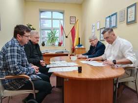 Pokaż zdjęcie: Przy owalnym stole siedzi czterech mężczyzn. Podpisują umowę. Na stole leżą różne dokumenty, pieczątki, długopis, klucze. 
