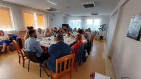 Pokaż zdjęcie: Kilkadziesiąt osób siedzi przy prostokątnym stole i rozmawia. Zwróceni są do siebie twarzami. Znajdują się w dużym pomieszczeniu. Na stole rozłożone są talerze, szklanki oraz kubki. Po lewej stronie zdjęcia uśmiechnięta kobieta siedząca na kanapie. 