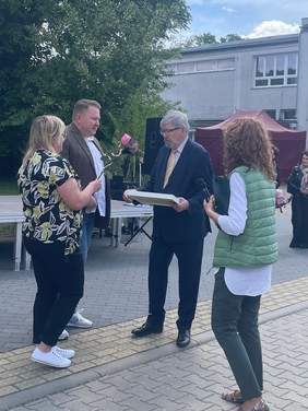 Pokaż zdjęcie: Na dziedzińcu stoją cztery osoby: dwie kobiety i dwóch mężczyzn. Jeden z panów odbiera dyplom za zaangażowanie i wsparcie. Jedna z pań trzyma różową różę. 