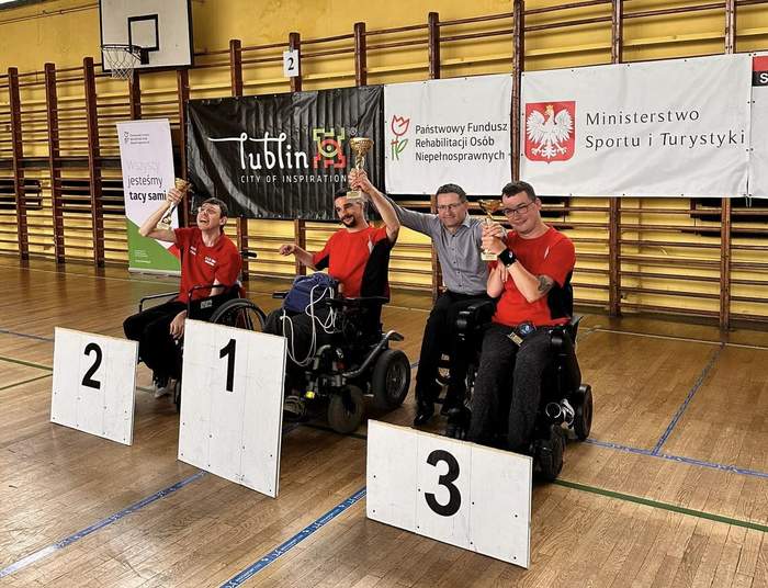 Pokaż zdjęcie: W sali sposrtowej na wózkach siedzi czterech mężczyzn, trzech z nich ma przed sobą stojące na podłodze tabliczki z numerem miejsca, które zajęli, pierwsze, drugie i trzecie.