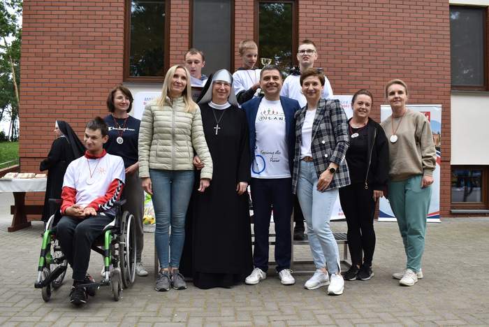 Pokaż zdjęcie: Do pamiątkowego zdjęcia na tle ośrodka  stanęli uczestnicy zawodów, wśród nich siostra zakonna, Prezydent Puław Paweł Maj, osoba siedząca na wózku inwalidzkim, pracownicy Oddziału Lubelskiego PFRON. 
