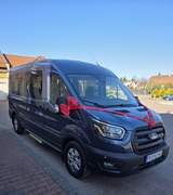 Ciemnoniebieski ośmioosobowy Ford z czerwoną wstążką i kwiatem na masce, zaparkowany na tle bezlistnych drzew.