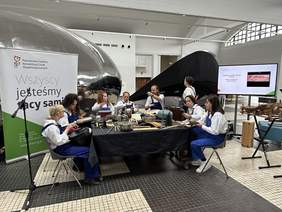 Pokaż zdjęcie: Przy kwadratowym stole siedzi osiem osób ubranych w granatowe ogrodniczki i białe koszule. Stół nakryty ciemnym obrusem, na nim leży wiele różnych instrumentów. Obok stoi telewizor z którego puszczana jest prezentacja na temat prac.