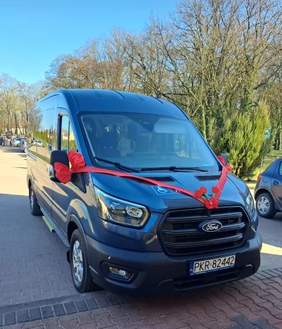 Pokaż zdjęcie: Ciemnoniebieski ośmioosobowy Ford z czerwoną wstążką i kwiatem na masce, zaparkowany na tle bezlistnych drzew.