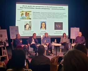 Pokaż zdjęcie: Panel dyskusyjny z pięcioma osobami siedzącymi na scenie przy stolikach z wodą; w tle slajd z informacjami i zdjęciami prelegentów; publiczność obserwuje wydarzenie.