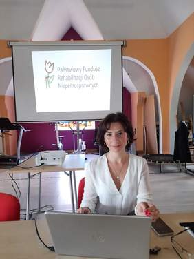 Pokaż zdjęcie: Za biurkiem siedzi młoda kobieta, w tle slajd z logo Państwowego Funduszu Rehabilitacji Osób Niepełnosprawnych.