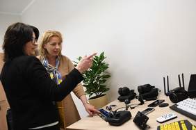 Pokaż zdjęcie: Pokój w którym stoi biurko, przy biurku rozmawiają dwie kobiety i oglądają położone na biurku sprzęty: linijka brajlowska, tablet, aparat, słuchawki, klawiatura