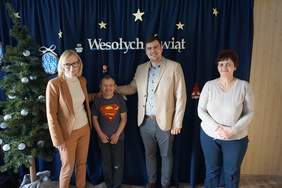 Pokaż zdjęcie: Przy świerku ozdobionym białymi lampkami i grantowej ścianie z napisem Wesołych świąt stoją: kierownik WTZ Karina Leliwa, uczestnik WTZ  w koszulce Supermena, pan dyrektor Mateusz Szkaradziński oraz wiceprezes Towarzystwa Przyjaciół Dzieci z Now