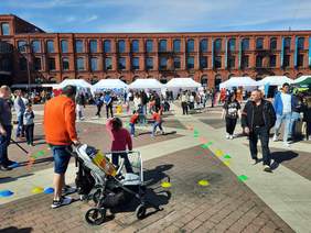Pokaż zdjęcie: Na rynku łódzkiej Manufaktury znajduje się tłum ludzi. Dzieci grają w piłkę, a dorośli i starsi spacerują. Jest piękna pogoda. 