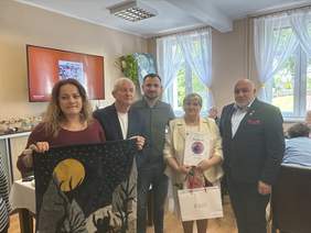Pokaż zdjęcie: Pięć osób stoi w eleganckim wnętrzu podczas uroczystego spotkania. Jedna osoba prezentuje ozdobną pracę przedstawiającą nocny krajobraz z księżycem, a inna trzyma dyplom i torbę z upominkiem. W tle widoczne są duże okna z dekoracyjnymi zasłonami oraz przygotowany poczęstunek. Zdjęcie upamiętnia wręczenie wyróżnienia połączone z prezentacją pracy artystycznej.