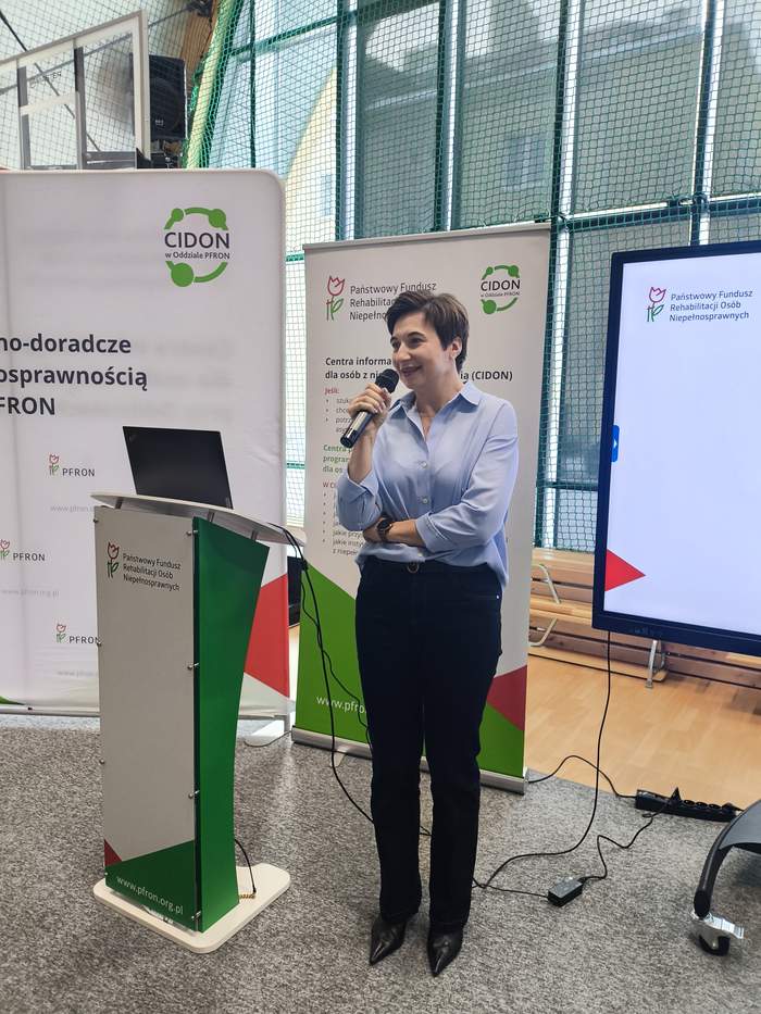Pokaż zdjęcie: Kobieta w niebieskiej bluzce prowadzi prezentację przy mównicy podczas wydarzenia PFRON. W tle stoją roll‑upy informacyjne.