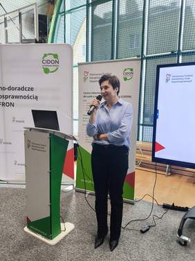 Pokaż zdjęcie: Kobieta w niebieskiej bluzce prowadzi prezentację przy mównicy podczas wydarzenia PFRON. W tle stoją roll‑upy informacyjne.