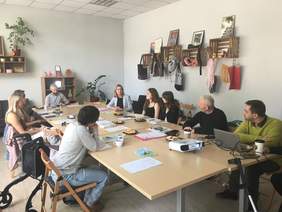 Pokaż zdjęcie: Jasny pokój z długim stołem, przy którym siedzi osiem osób w różnym wieku, w trakcie spotkania lub warsztatów, z laptopami, projektorem i przekąskami na stole.