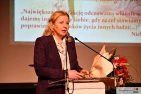 Pokaż zdjęcie: Przy mównicy stoi kobieta blondynka ubrana w ciemny garnitur, na mównicy leżą kwiaty.