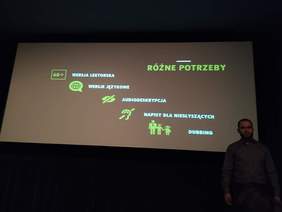 Pokaż zdjęcie: Sala w Kinie pod Baranami. Na ekrane informacje dotyczące potrzeb osób z niepełnosprawnościami i aplikacji AudioMovie. Przed ekranem stoi Piotr Franiek.
