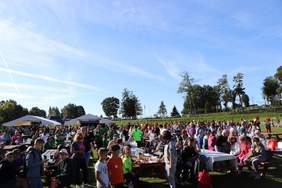 Pokaż zdjęcie: Na polu stoi wiele długich ław przy których siedzą lub chodzą ludzie. W tle namioty estradowe i drzewa.