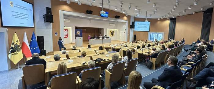 Pokaż zdjęcie: Na dużej Sali wykładowej przy drewnianych biurkach ułożonych w półokręgu siedzą ludzie. Naprzeciw nich za mównicą stoi kobieta w blond włosach ubrana w szarą marynarkę. Po prawej stronie wisi telebim.