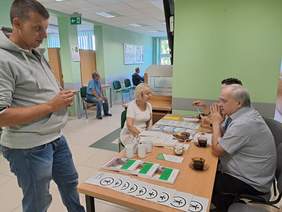 Pokaż zdjęcie: Przy biurku na którym leżą katalogi i ulotki siedzą trzy osoby, dwie kobiety i mężczyzna. Kobiety rozmawiają ze sobą. Koło biurka stoi mężczyzna w bluzie dresowej.