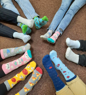 Pokaż zdjęcie: Na wykładzinie siedzą ludzie. Nogi ułożyli w kółko. Wszyscy mają założone kolorowe skarpetki. 