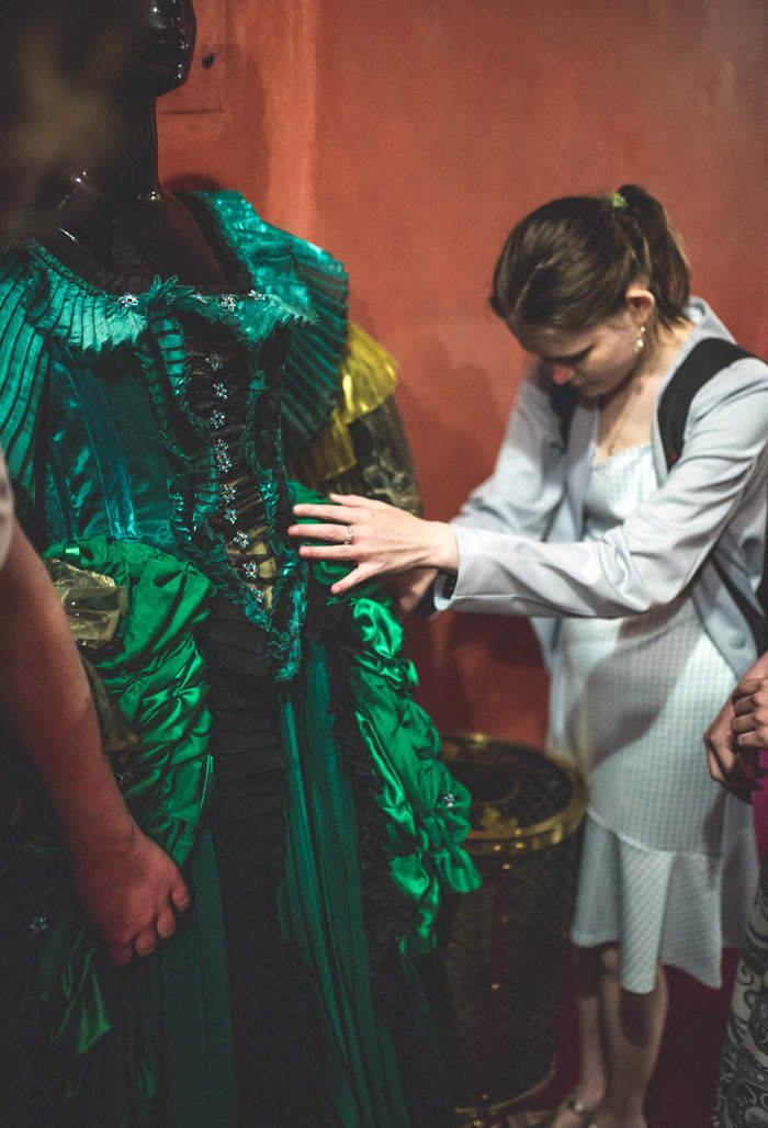 Pokaż zdjęcie: Niewidoma dziewczyna dotyka zieloną, bogato zdobioną suknię, która jest kostiumem teatralnym. Suknia jest na manekinie.