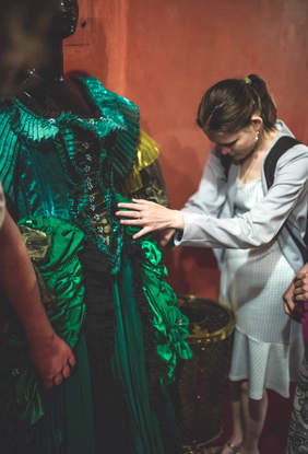 Pokaż zdjęcie: Niewidoma dziewczyna dotyka zieloną, bogato zdobioną suknię, która jest kostiumem teatralnym. Suknia jest na manekinie.