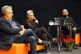 Pokaż zdjęcie: Trzy osoby siedzące na scenie z mikrofonami w ręku