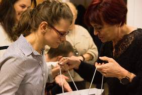 Pokaż zdjęcie: Dwie kobiety stoją na przeciw siebie starsza w dłoniach trzyma otwartą torbę prezentową, obie zaglądają do środka torby.