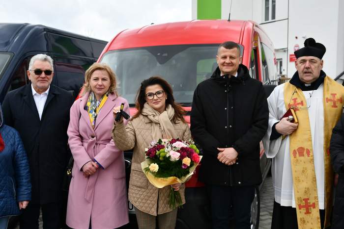 Pokaż zdjęcie: Do pamiątkowego zdjęcia na tle czerwonego busa pozuje 5 osób, od lewej mężczyzna w ciemnych okularach, kobieta blondynka, kobieta trzymająca bukiet, mężczyzna w płaszczu oraz ksiądz.