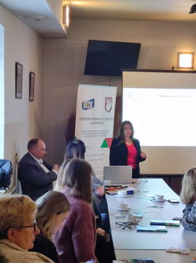 Pokaż zdjęcie: Z tyłu sali w kierunku prelegentki zrobiono zdjęcie pokazujące uczestników siedzących przy długom stole. Za prelegentką stoi ekran oraz rollupy.