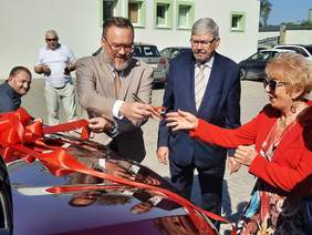 Pokaż zdjęcie: Przy samochodzie stoi pani i dwóch panów, którzy przecinają wstęgę położoną na samochodzie.