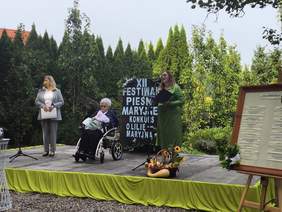 Pokaż zdjęcie: Na scenie są trzy kobiety, w tym jedna na wózku inwalidzkim. Trwa zapowiedź występu wokalnego.