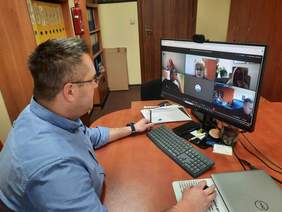 Pokaż zdjęcie: Trwa wideokonferencja. Na ekranie monitora widać różne osoby uczestniczące w spotkaniu. Przed komputerem siedzi mężczyzna. 