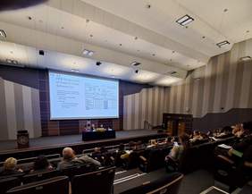 Pokaż zdjęcie: Sala wykładowa z audytorium i dużym ekranem projekcyjnym na scenie. W kadrze widać rzędy siedzeń z ludźmi siedzącymi i słuchającymi, a na dole pośrodku jest scena z długim stołem, przy którym siedzą trzy osoby. Na ekranie wyświetlony jest slajd z dużą ilością tekstu i tabelą. Pomieszczenie ma wysokie, jasne sufity z prostokątnymi lampami oraz ściany w pionowe pasy w odcieniach beżu i szarości.