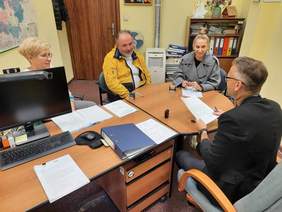 Pokaż zdjęcie: Przy stole siedzą cztery osoby: dwie kobiety i dwóch mężczyzn. Rozmawiają ze sobą i patrzą na siebie. Na stole leżą różne dokumenty i segregatory. 