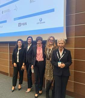 Pokaż zdjęcie: Zdjęcie grupowe pięciu kobiet stojących w sali konferencyjnej przed dużym ekranem projekcyjnym. Kobiety stoją obok siebie i są ubrane oficjalnie. W tle widać drewniane panele ścienne z poziomymi jasnymi liniami oraz fragment dużego slajdu na ekranie. Na slajdzie są logotypy partnerów Konferencji szkoleniowo-edukacyjnej pod tytułem: Psychoterapia młodych. 