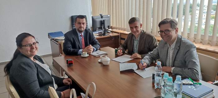 Pokaż zdjęcie: Przy stoliku podpisują umowę przedstawiciele Powiatu i PFRON - trzej mężczyźni i jedna kobieta, wszyscy są uśmiechnięci i ubrani na galowo