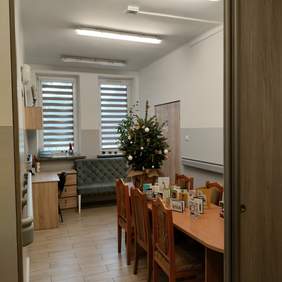 Pokaż zdjęcie: Zdjęcie pokoju w którym znajduje się zielona sofa do siedzenia, po lewej stronie stoi biurko w kolorze drzewa, nad nim wisi szafka. Po prawej stronie zdjęcia widać duży stół z krzesłami w kolorze olchy. Na stole stoi udekorowana świątecznie choinka. Na ścianach zamontowane są uchwyty.