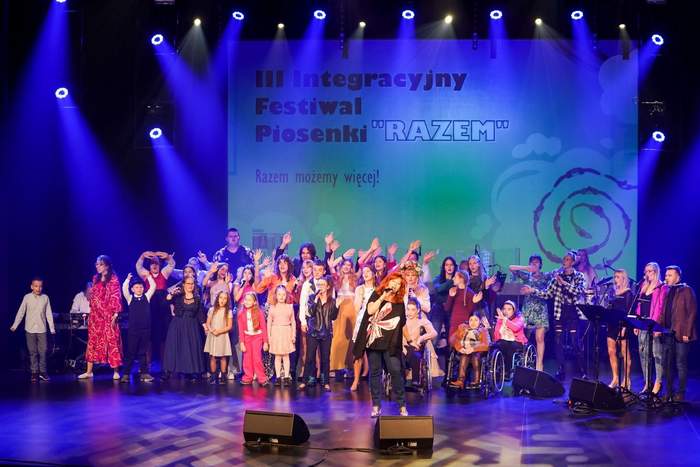 Pokaż zdjęcie: Wspólny występ na scenie dużej grupy osób, zarówno dzieci, młodzieży, jak i dorosłych. Większa część osób ma uniesione w górę ręce. Artyści oświetleni są niebieskimi światłami. W tle widoczny jest duży ekran, na którym wyświetlone są następujące sformułowania: III Integracyjny Festiwal Piosenki „Razem” oraz „Razem możemy więcej!”. Przed wykonawcami możemy dostrzec również cztery ustawione na scenie odsłuchy.