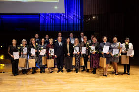 Pokaż zdjęcie: Na scenie Filharmonii Łódzkiej stoją elegancko ubrani ludzie. Trzymają w dłoniach róże, dyplomy z podziękowaniami i torby prezentowe. 