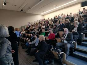 Pokaż zdjęcie: W sali konferencyjnej na widowni siedzą w rzędach ludzie. Są to słuchacze semunarium. Przysłuchują się wykładom.