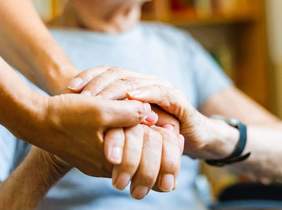 Pokaż zdjęcie: Grafika z widocznymi dłońmi młodej osoby, opiekuna, trzymającymi dłonie starszej osoby. 