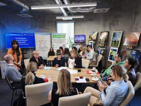 Pokaż zdjęcie: Spotkanie grupy kilkunastu osób przy dużym stole w nowoczesnej sali konferencyjnej. Uczestnicy rozmawiają i przeglądają dokumenty. W tle widoczne są plansze z informacjami o wsparciu dla lokalnych organizacji pozarządowych oraz wystawa fotografii ustawiona na sztalugach.