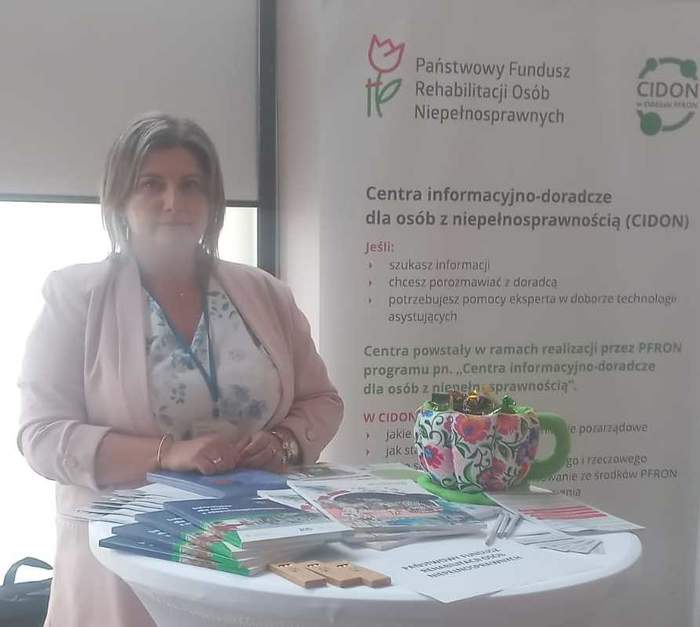 Pokaż zdjęcie: Przy okrągłym stoliku na którym leżą ulotki stoi kobieta w jasnoróżowej marynarce, w tle rollup z logo CIDON oraz Funduszu.