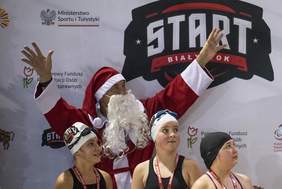 Pokaż zdjęcie: Osoba przebrana za Świętego Mikołaja, z uniesionymi rękoma, stoi na tle baneru z logo „START Białystok” oraz logotypami Ministerstwa Sportu i Turystyki, PFRON i innych partnerów. Przed nim stoją trzy młode zawodniczki w czepkach pływackich, z medalami na szyjach.