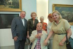 Pokaż zdjęcie: Zdjęcie sześciu osób, od lewej stoi ksiądz w cywilnym ubraniu, szczupła blondynka, obok starsza kobieta, kobieta z rudymi włosami oraz blondynka w beżowej sukience, która trzyma za rękę mężczyznę siedzącego na wózku. Zdjęcie w muzem, w tle widać obraz.