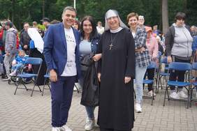 Pokaż zdjęcie: Zdjęcie czterech osób, mężczyzna w garniturze, kobieta z ciemnymi włosami, siostra zakonna i kobieta z krótkimi włosami w marynarce w kratkę. W tle widać uczestników paraolimpiady