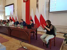 Grupa kilku osób, kobiet i mężczyzn przy stole prezydialnym, za nimi flagi biało-czerwone