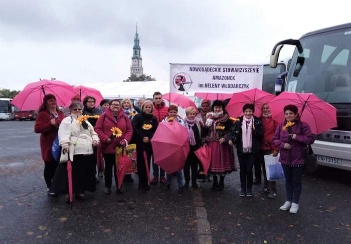 Pokaż zdjęcie: Na zdjęciu Zarząd i członkinie Nowosądeckiego Stowarzyszenia Amazonek - uśmiechnięte panie stoją w pochmurny dzień na parkingu, w rękach trzymają żółte kwiaty i rozłożone różowe parasole.
