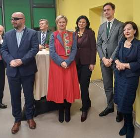 Pokaż zdjęcie: Grupa sześciu elegancko ubranych osób stoi w jasnym, żółtym wnętrzu podczas formalnego spotkania. W tle widoczny wysoki stolik koktajlowy i okno.