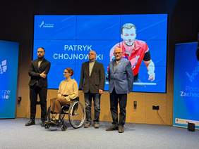 Pokaż zdjęcie: Zdjęcie przedstawia cztery osoby stojące na scenie na tle dużego ekranu. Po lewej stronie młody mężczyzna w czarnym garniturze i golfie, obok niego kobieta w okularach i jasnej bluzce, siedząca na wózku inwalidzkim. Dalej stoją dwaj starsi mężczyźni: jeden w ciemnej marynarce i szarym golfie, drugi w niebieskiej marynarce. Na ekranie w tle widnieje napis „PATRYK CHOJNOWSKI” oraz zdjęcie sportowca grającego w tenisa stołowego. Po lewej stronie widać fragment baneru z napisem „Pomorze Zachodnie”.